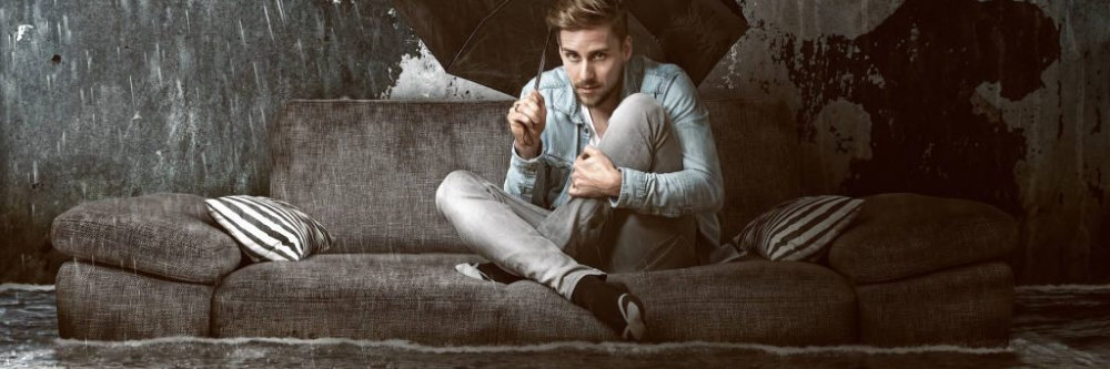 Man with an umbrella sitting on a sofa wondering what would happen if his flooded carpet was left untreated